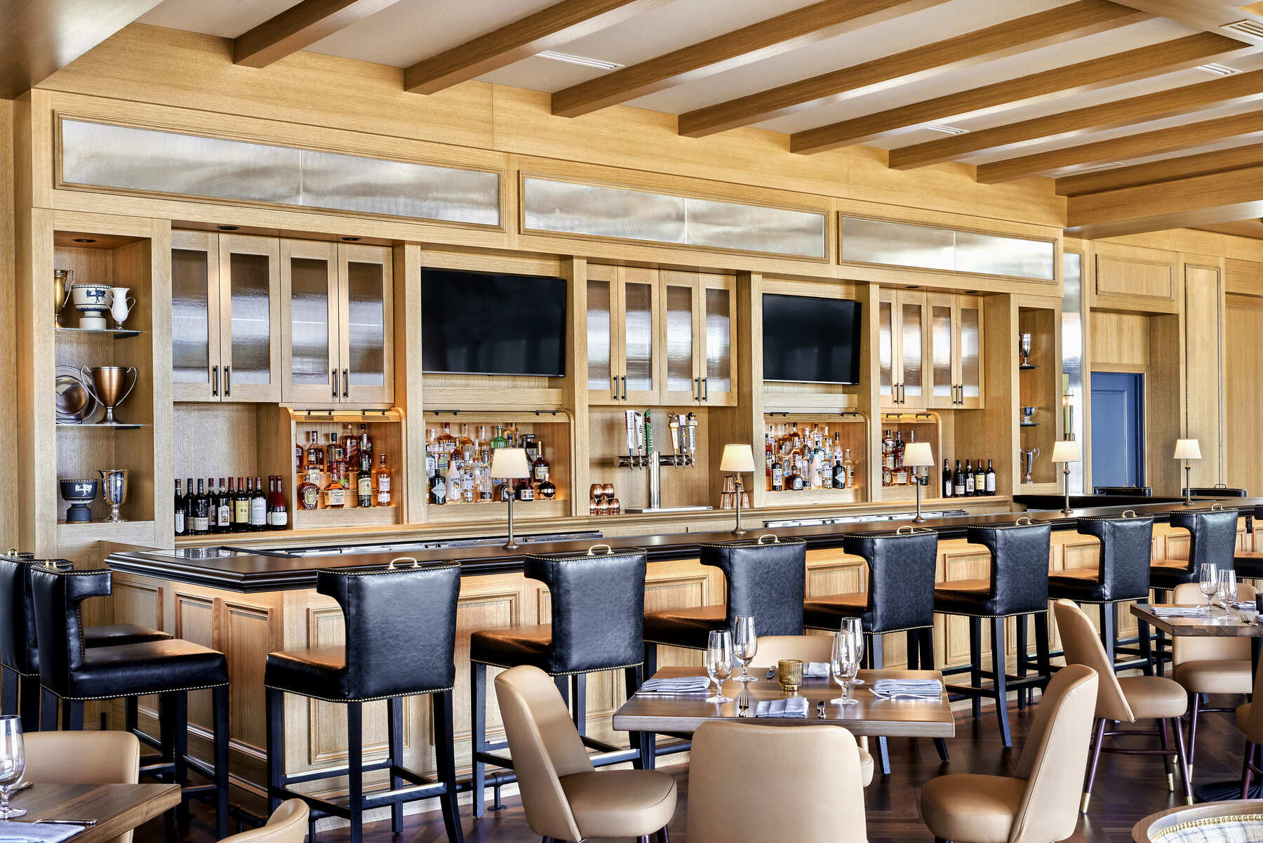 The modern bar area of the Ryder Cup Grille at Omni PGA Frisco, featuring light wood finishes, bar seating, and liquor shelves with inset televisions.
