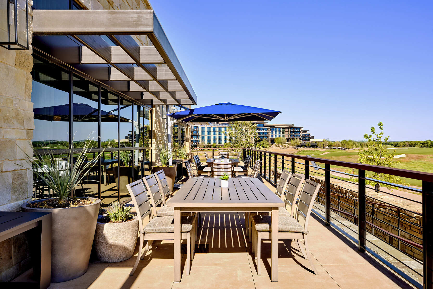 Outdoor patio dining area with a long table, blue umbrellas, and a view overlooking the green golf course.