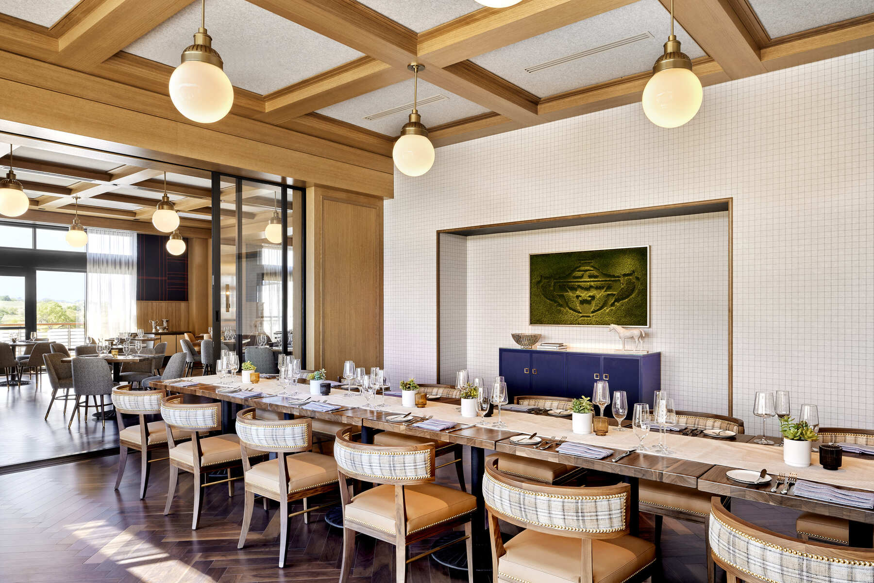 A glass-enclosed private dining room with a long wooden table, spherical lights, and green artwork on a white tiled wall.