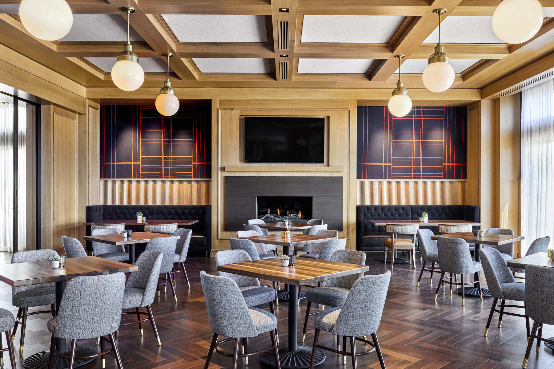 Spacious restaurant interior featuring wood coffered ceilings, globe pendant lights, and modern gray seating around wooden tables.