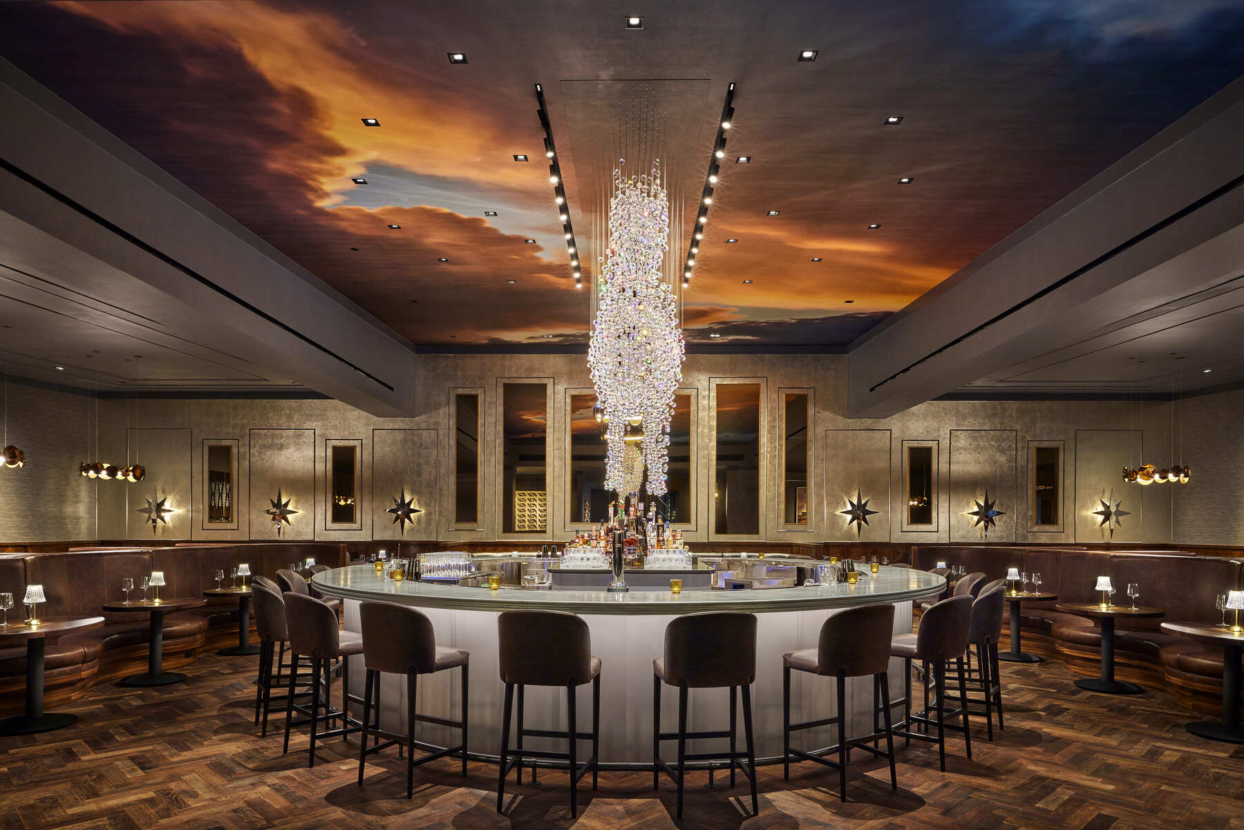 The luxurious circular central bar at Trick Rider restaurant, featuring a dramatic crystal chandelier and a ceiling mural resembling a sunset sky.