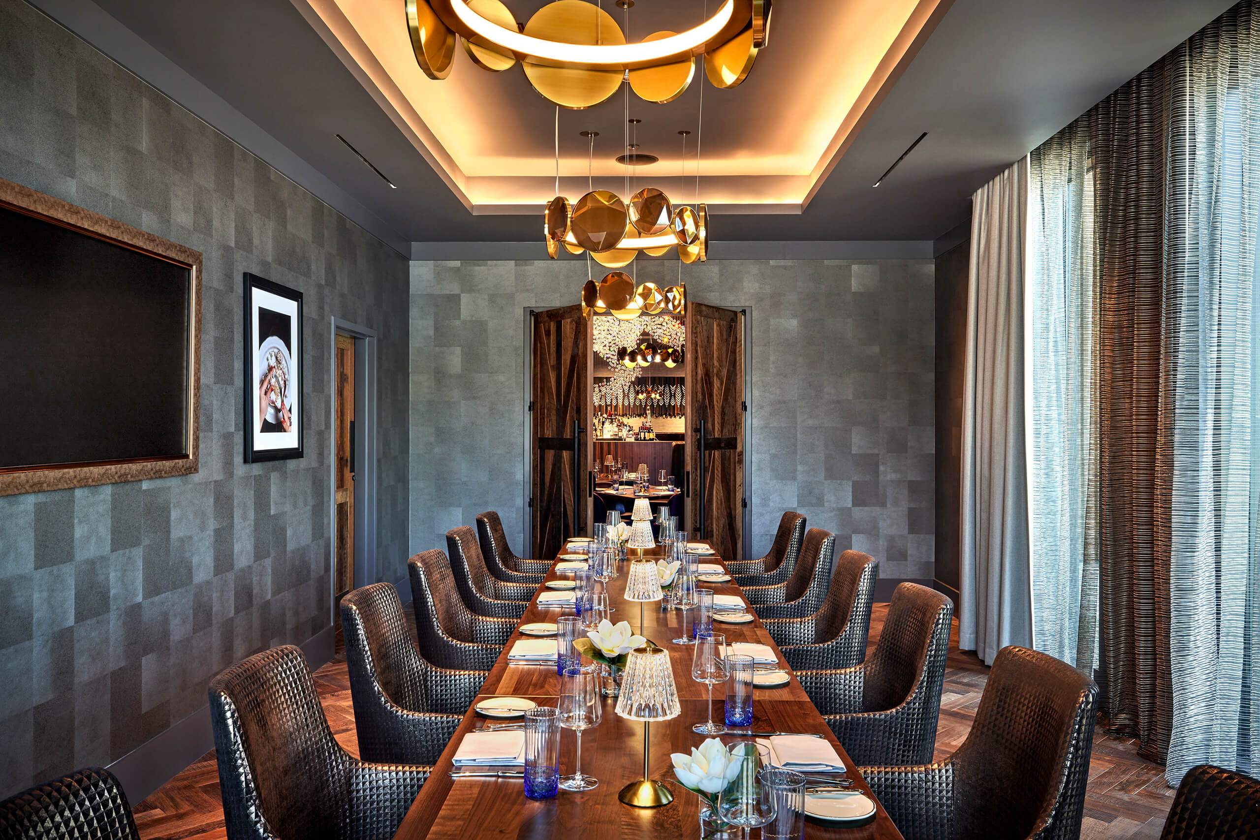 An elegant private dining room featuring a long wooden table set for dinner, textured grey walls, and a modern gold ring chandelier.