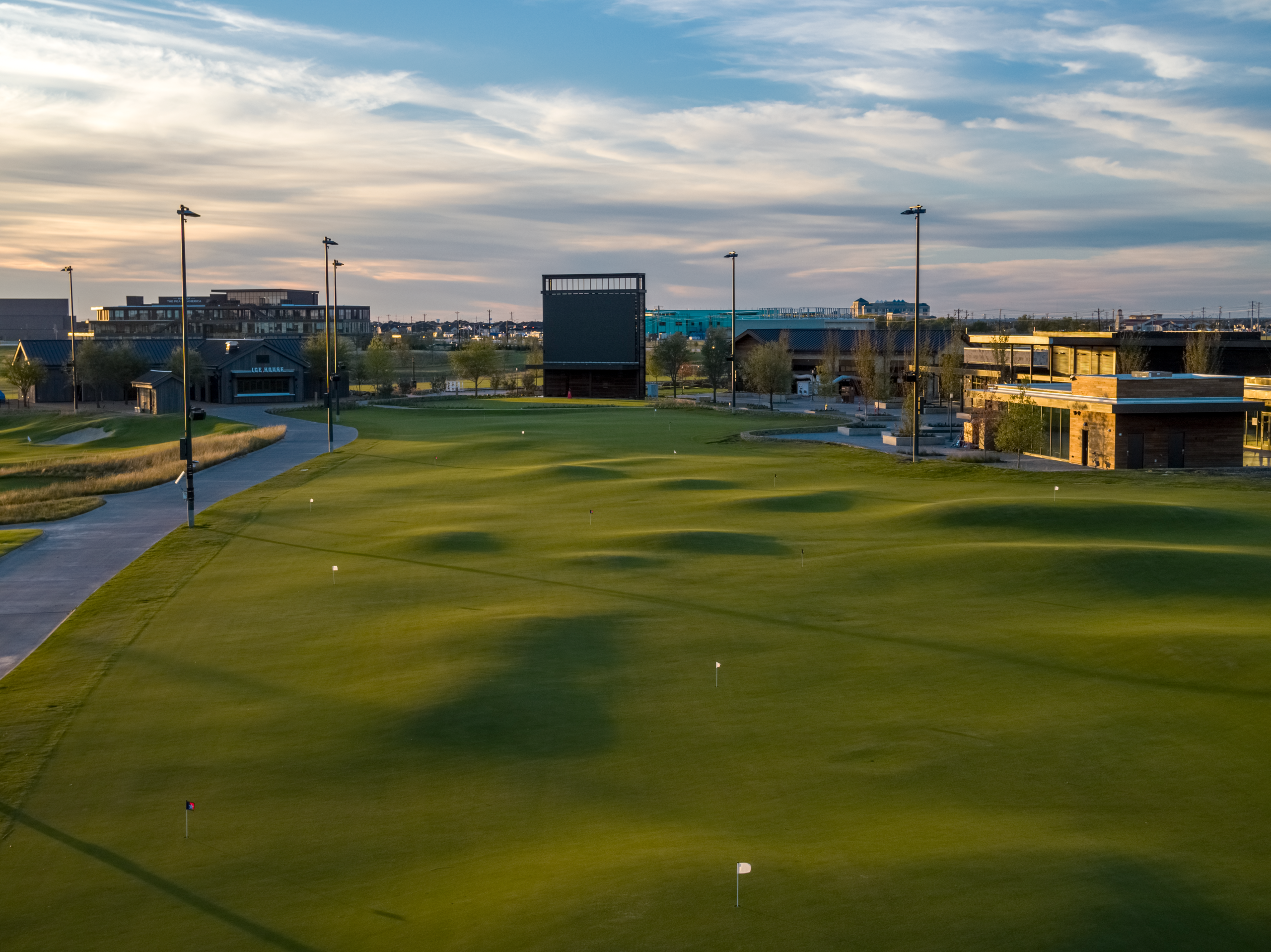 A large, undulating putting green with multiple flagsticks, surrounded by buildings and a giant video scoreboard at dusk.