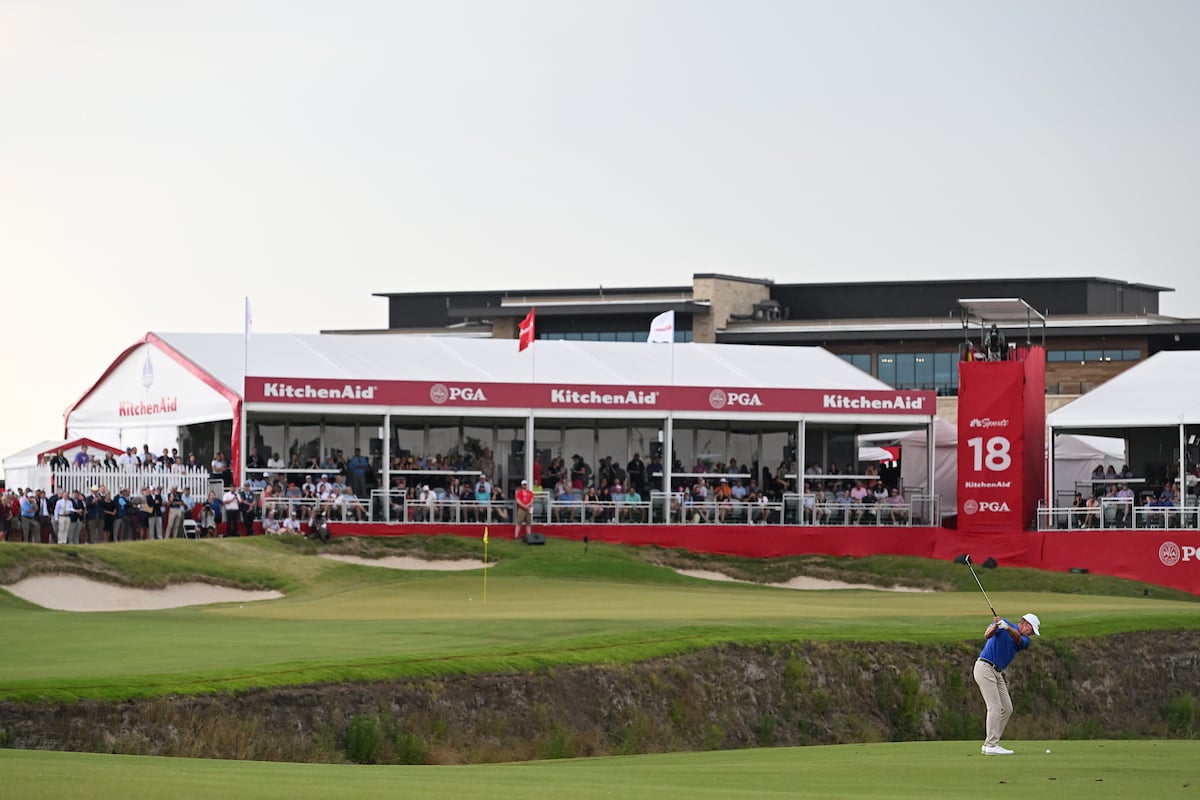 A golfer is mid-swing on the fairway near the 18th hole, with a large, red 
