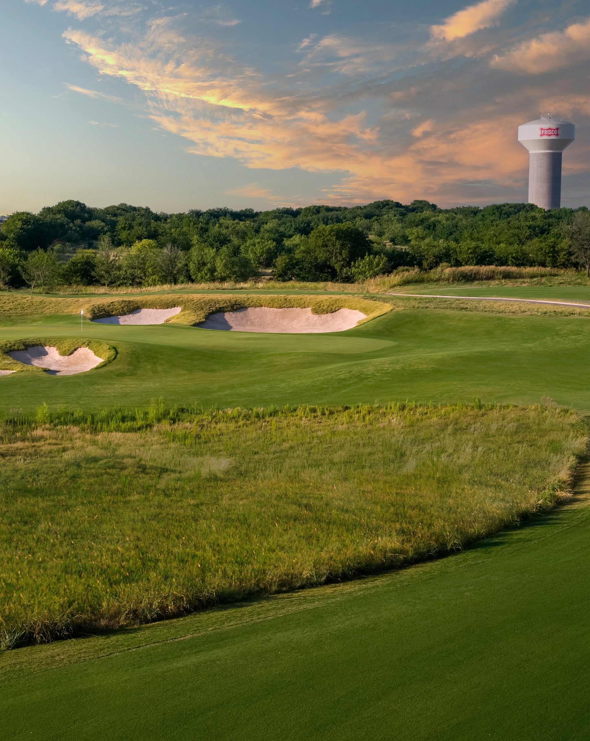 A green golf hole with sand bunkers and native grasses, backed by a treeline and a white water tower at sunset.