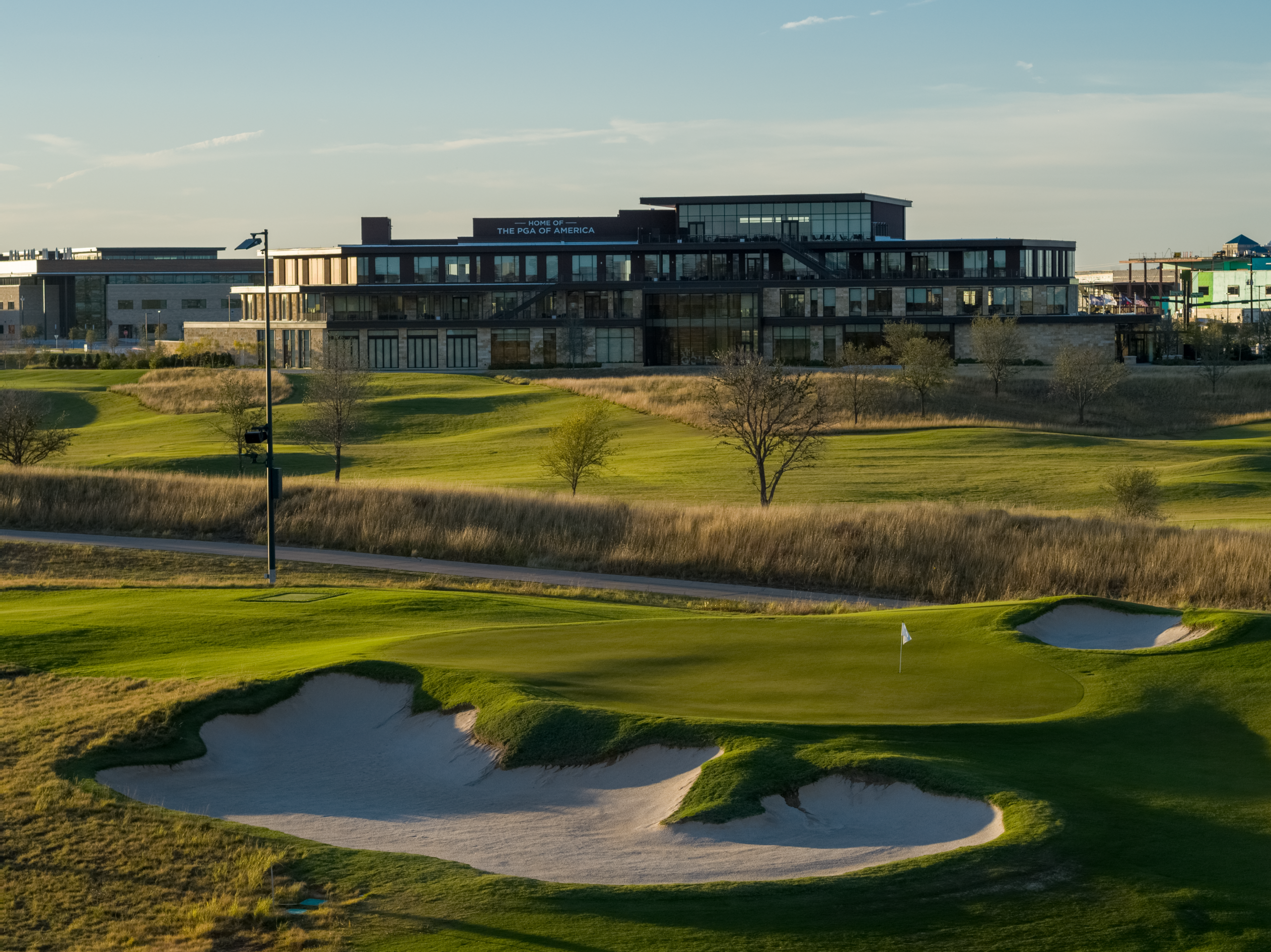 A golf green and sand trap in the foreground, with the modern, glass-paneled PGA of America headquarters building in the background.
