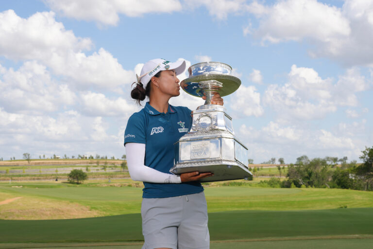 Female golfer Minjee Lee kisses a large, silver PGA Championship trophy on a green course under a cloudy blue sky.