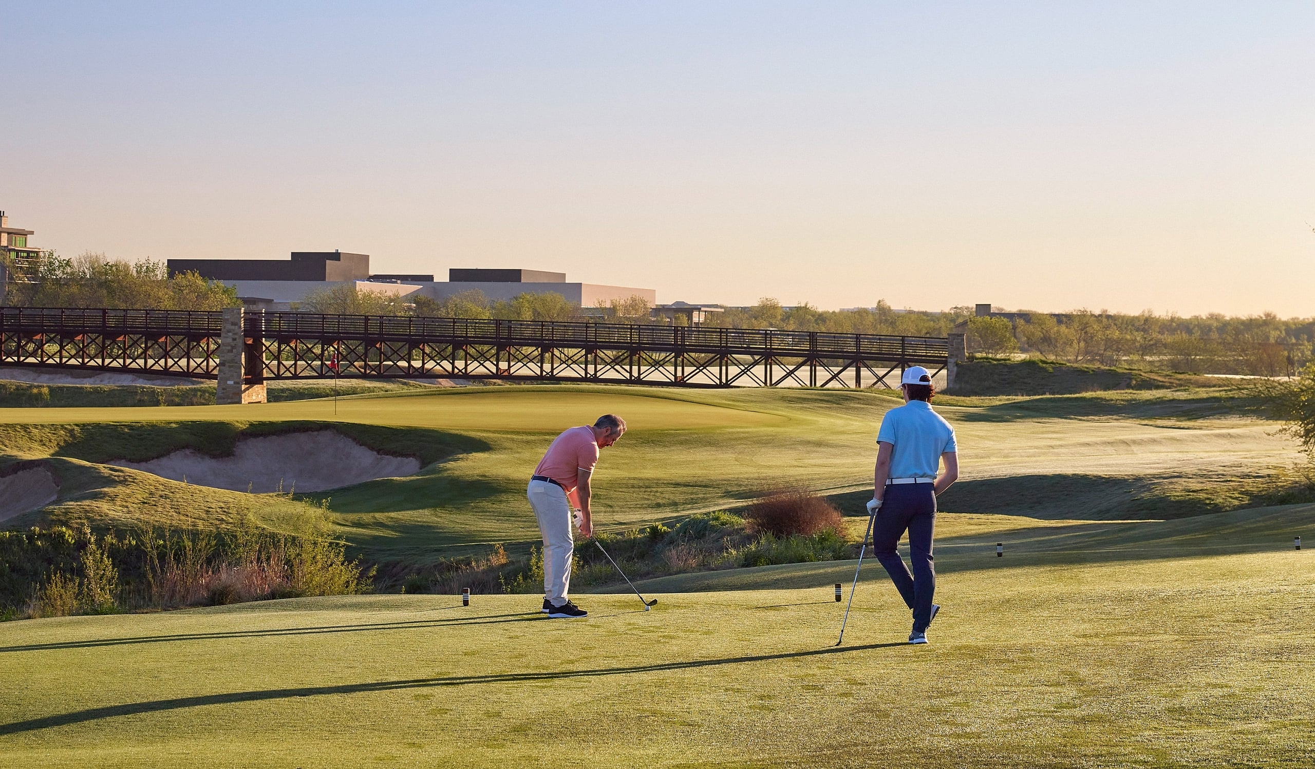 Two golfers putting on a green during golden hour with a bridge and resort buildings in the distance.