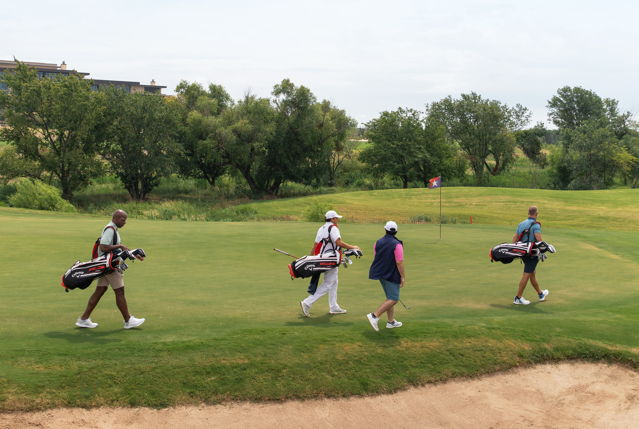 Four golfers carrying bags walking single-file across a fairway towards a Texas-themed flag.