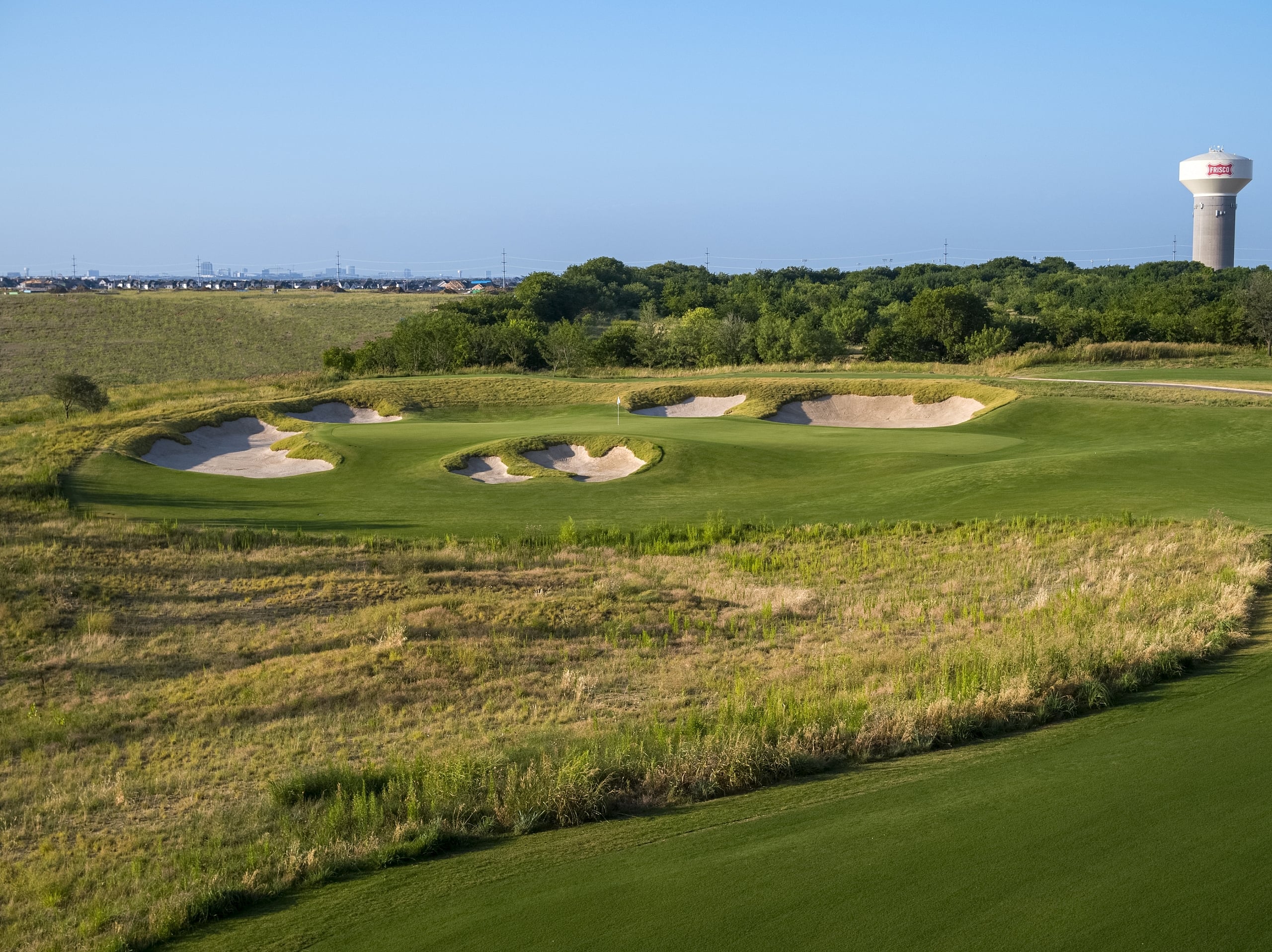 View of a golf course hole with large, strategically placed sand bunkers near the green, set against a distant treeline and a water tower.