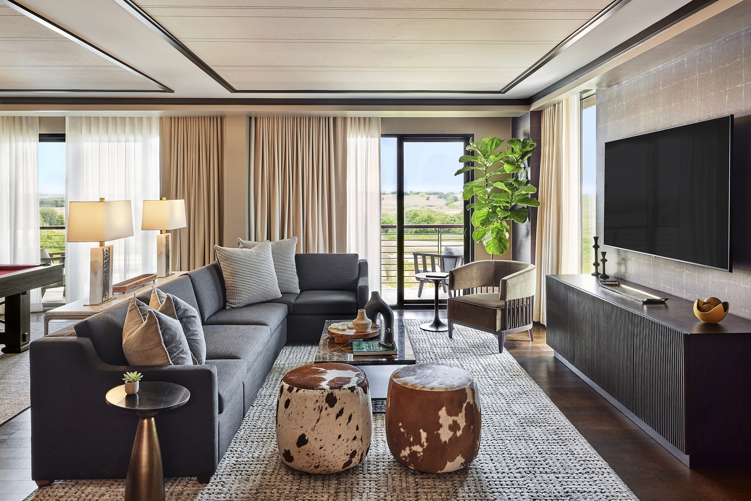Luxurious suite living room with a dark sectional sofa, cowhide ottomans, a patterned rug, and a glass door leading to a balcony.