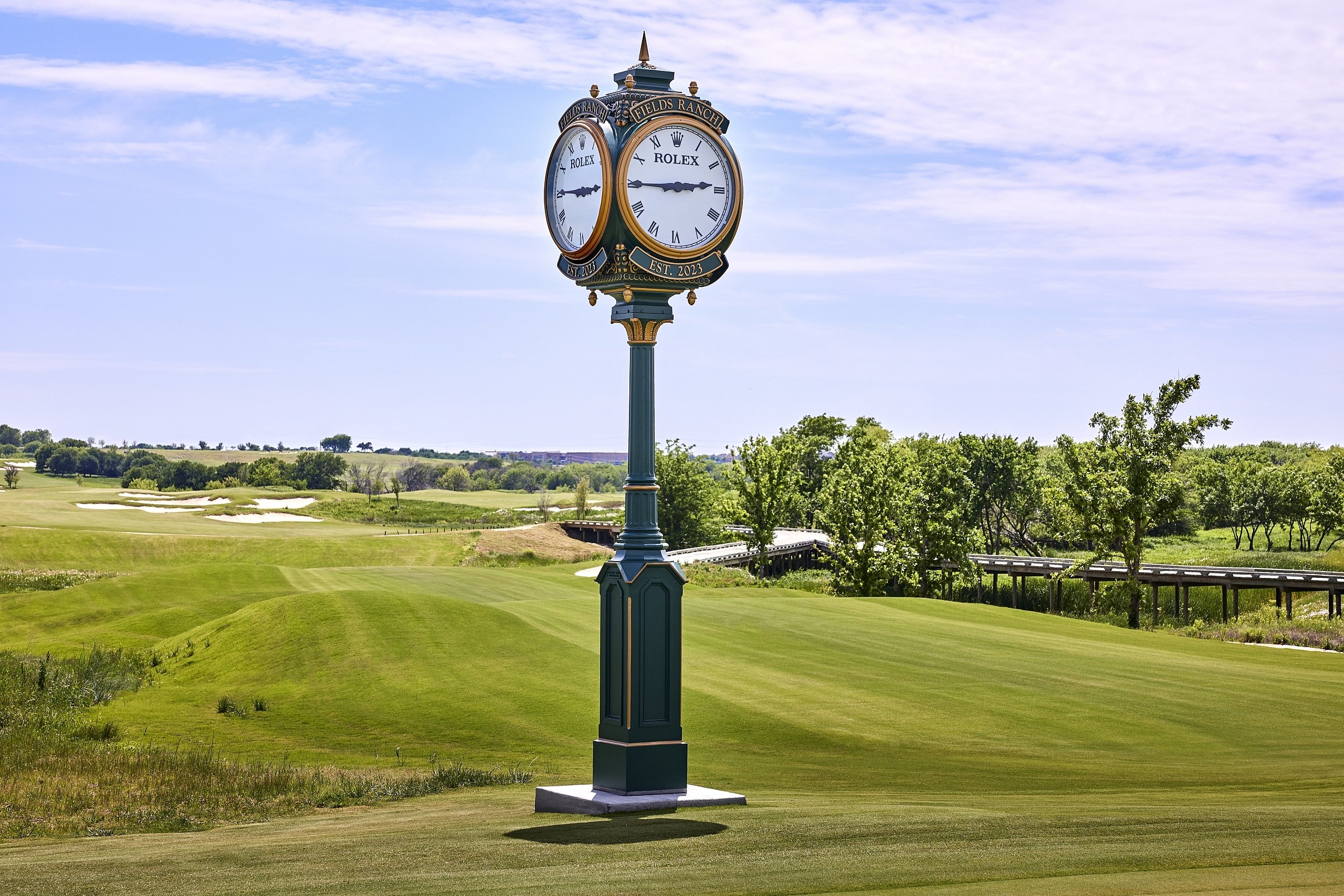 a rolex clock tower stands in front of a fairway on a sunny day