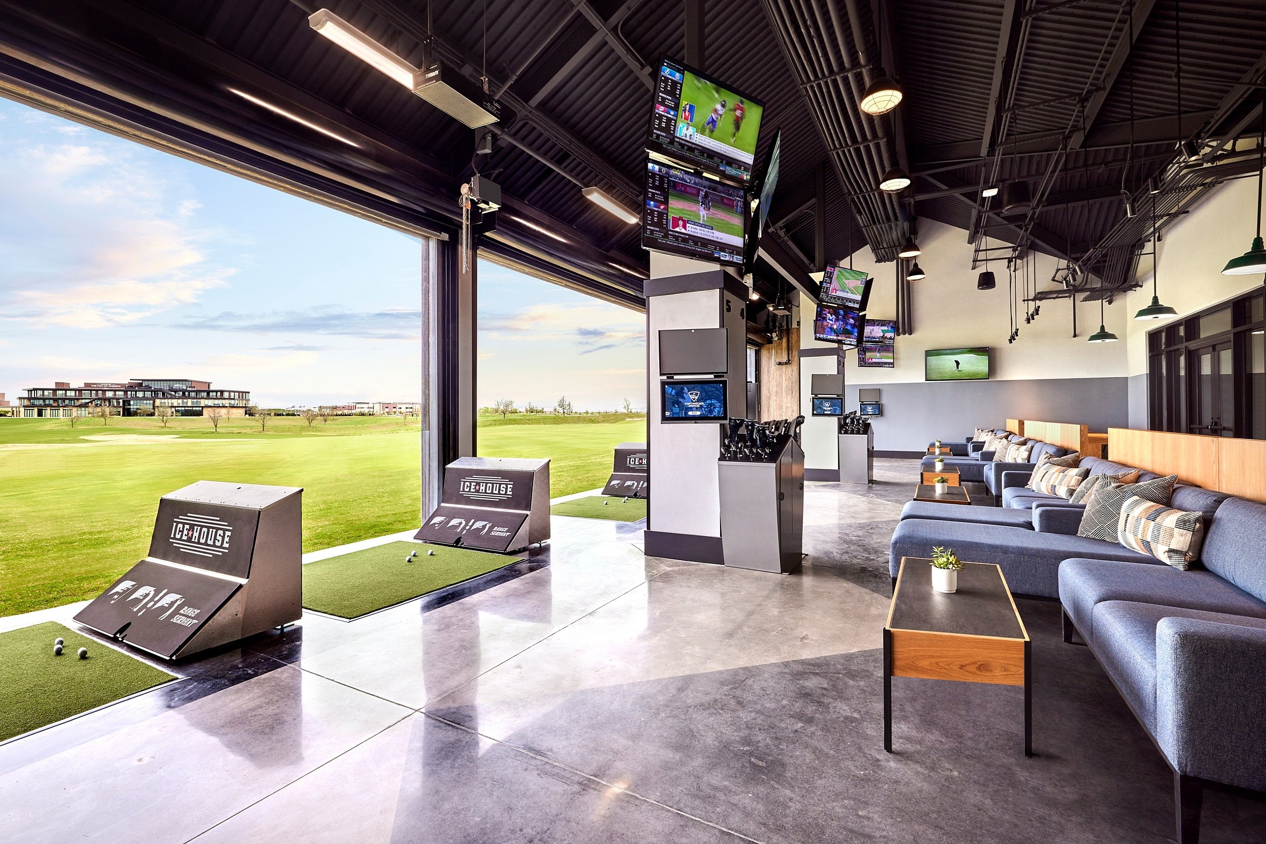 Indoor-outdoor golf hitting bays with artificial turf, seating areas, and multiple TV screens showing sports, overlooking a wide green golf course outdoors.