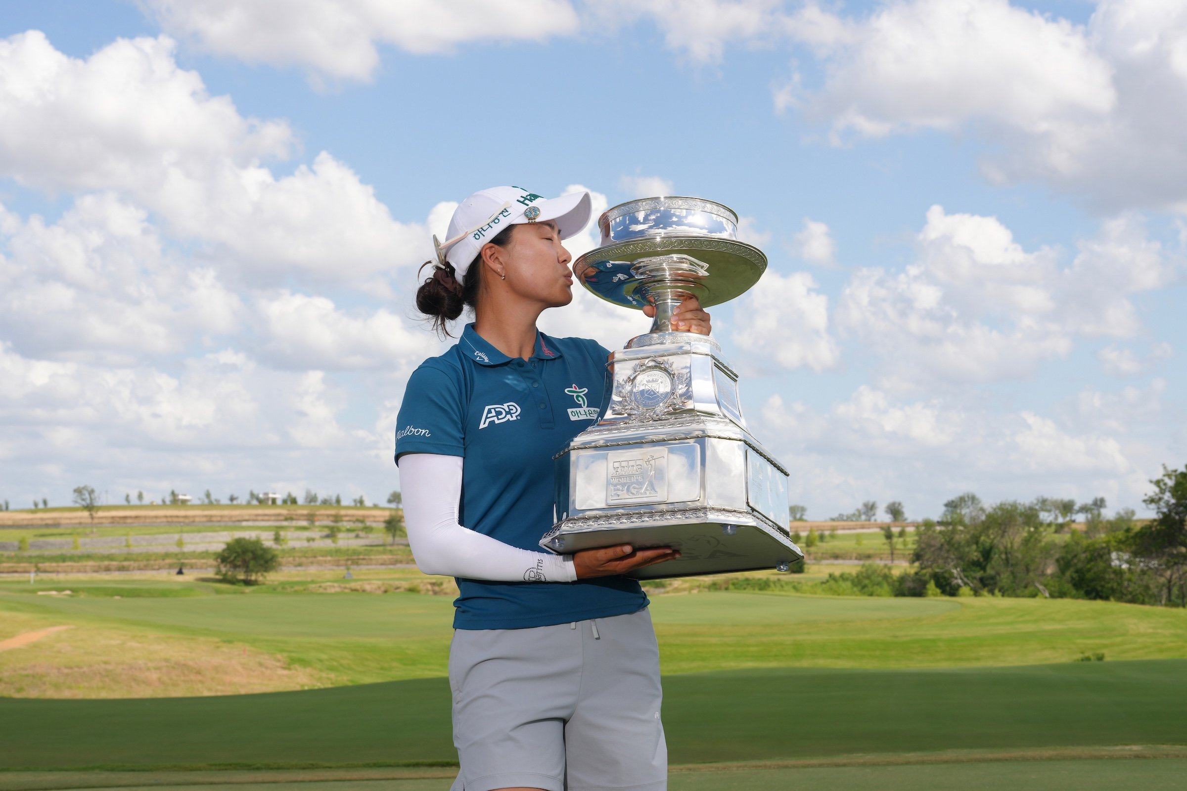 Female golfer Minjee Lee kisses a large, silver PGA Championship trophy on a green course under a cloudy blue sky.