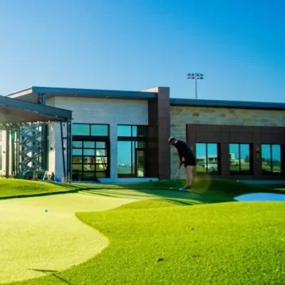 5. NTX-PGA-Ronny-DSC_5869...webp A golfer putting on a sunny green artificial turf practice area outside a modern clubhouse building.