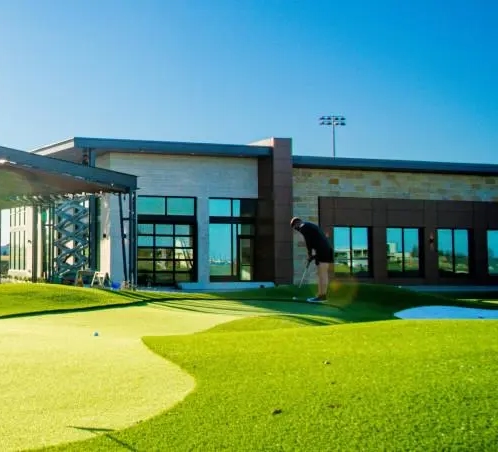 5. NTX-PGA-Ronny-DSC_5869...webp A golfer putting on a sunny green artificial turf practice area outside a modern clubhouse building.