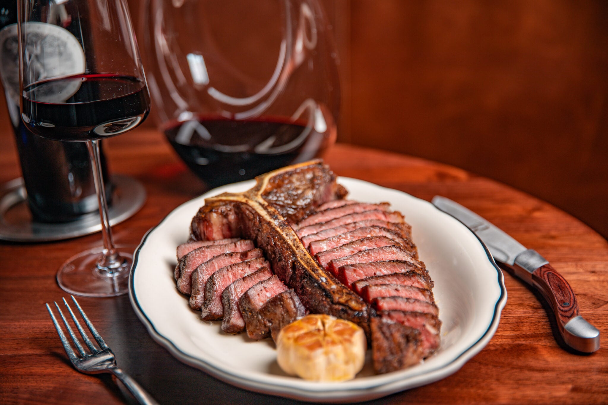 A sliced, perfectly cooked T-bone steak served on a white plate with roasted garlic and a glass of red wine.