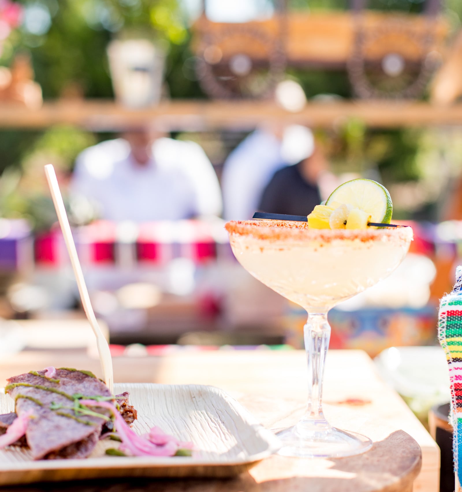 A fresh margarita with a chili-salted rim and lime garnish next to a plate of tacos at an outdoor event.