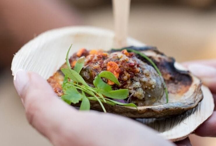 A hand holding a small wooden bowl containing a gourmet grilled oyster topped with herbs and relish.