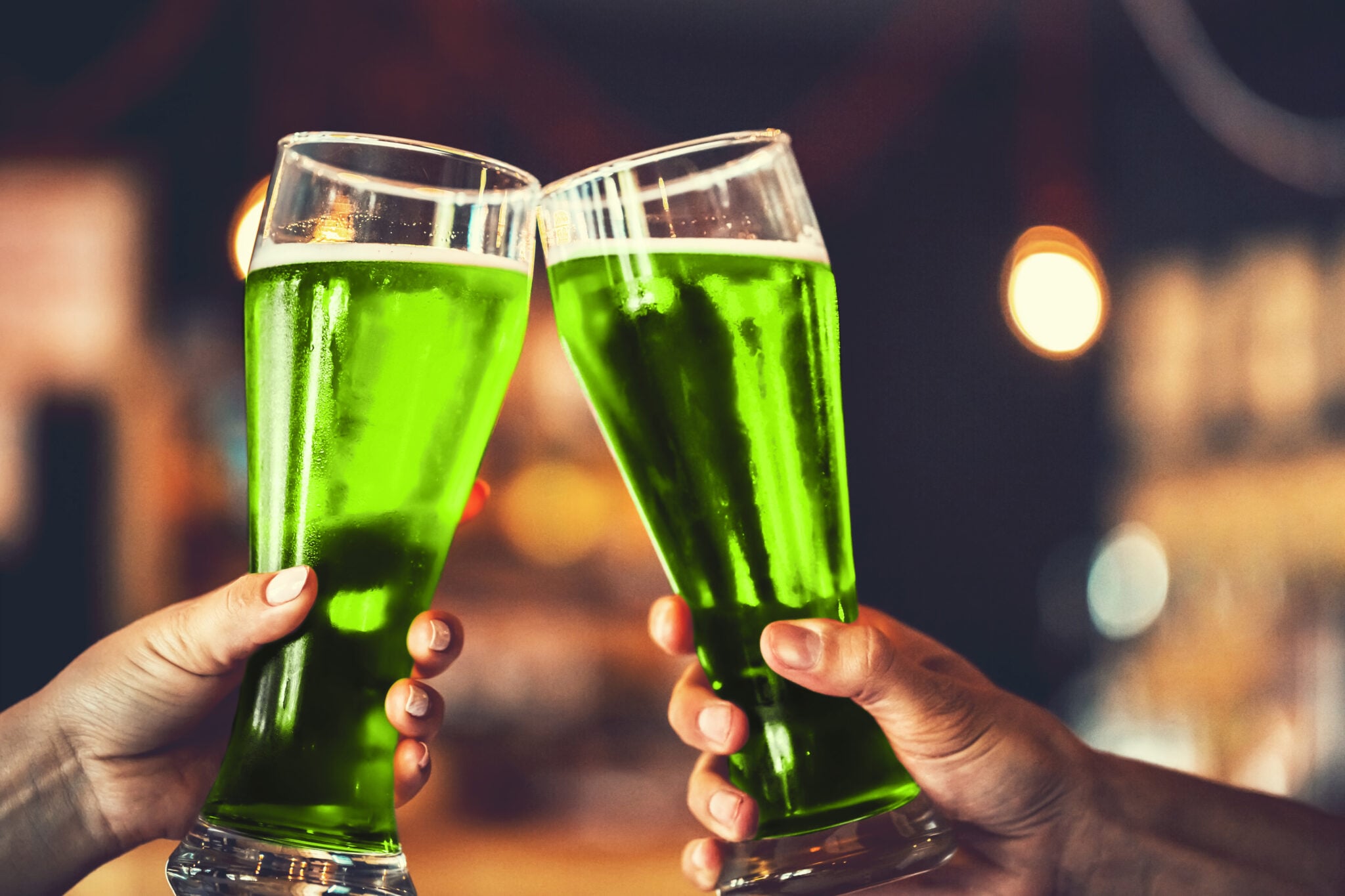 Two hands clinking tall glasses filled with green beer in a celebratory toast at a dimly lit bar.