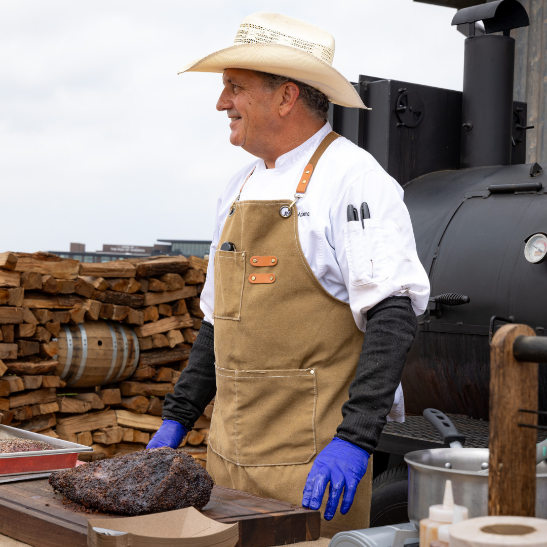 A smiling chef in a cowboy hat and apron standing next to a large black smoker and a seasoned brisket.