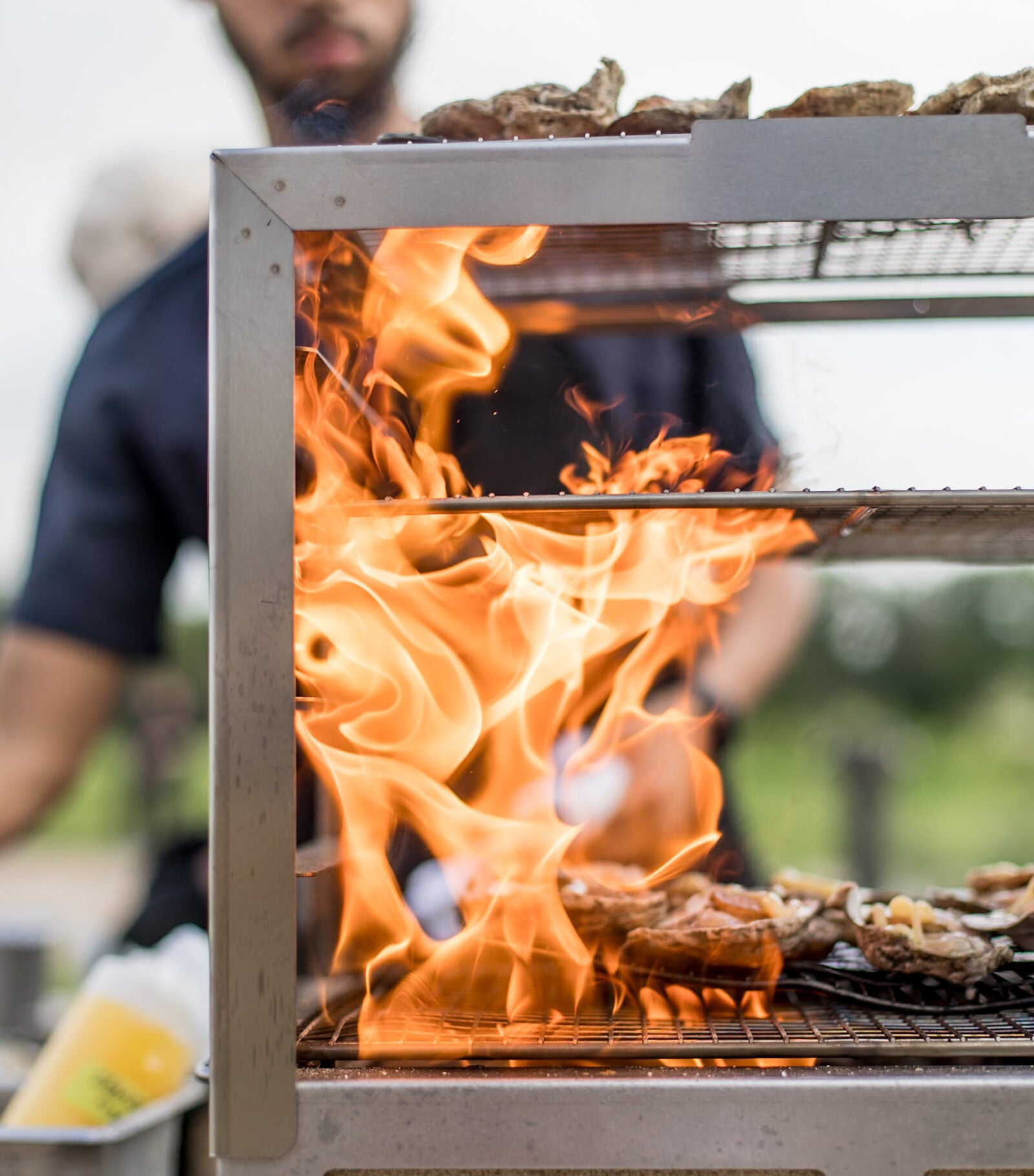 Bright orange flames leaping from an outdoor grill as a chef prepares food in the background.